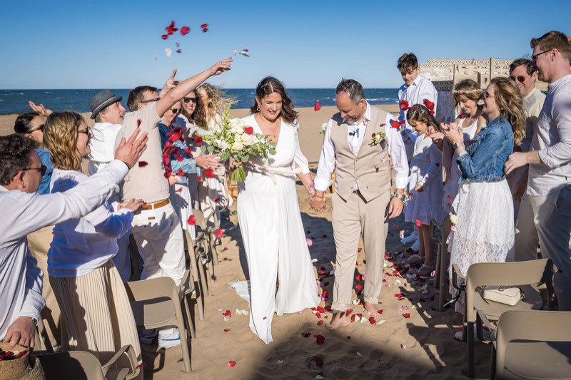 Los novios salen de la ceremonia en la playa entre pétalos y los invitados